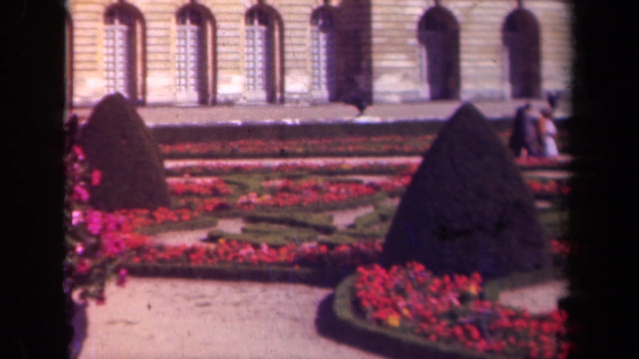 1939 France home-movie frame showing palace-style gardens with red flowers, dark conical hedges, and arched windows behind them.