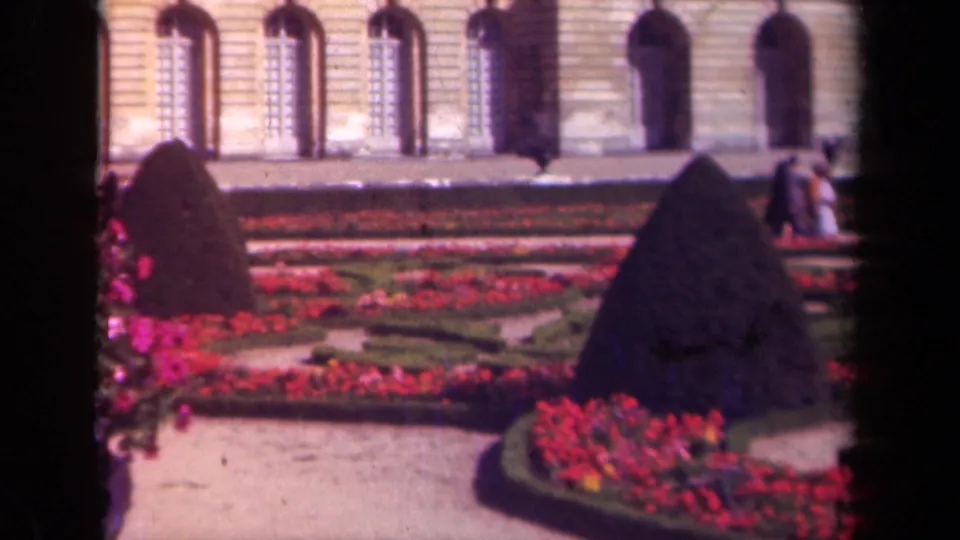 1939 France home-movie frame showing palace-style gardens with red flowers, dark conical hedges, and arched windows behind them.
