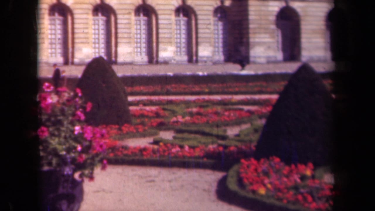 1939 France frame with formal garden beds, clipped hedges, and visitors walking near a palace-like stone facade.