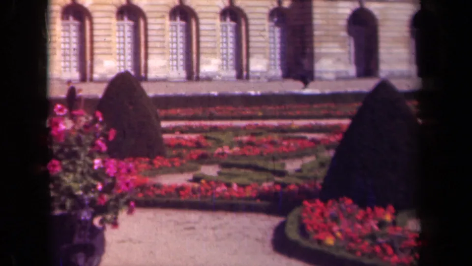 1939 France frame with formal garden beds, clipped hedges, and visitors walking near a palace-like stone facade.