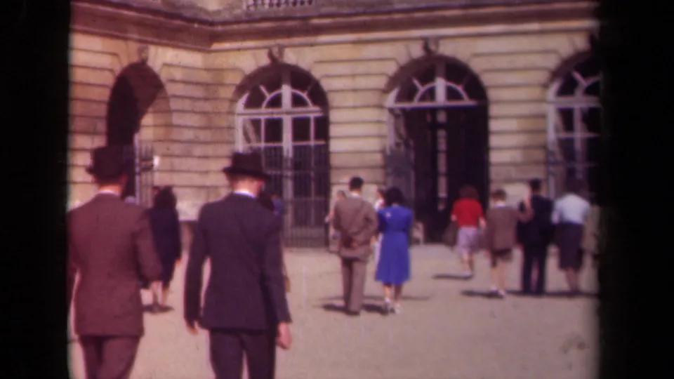 1939 France home-movie frame showing two formally dressed men in top hats walking toward palace-style arched windows and visitors.