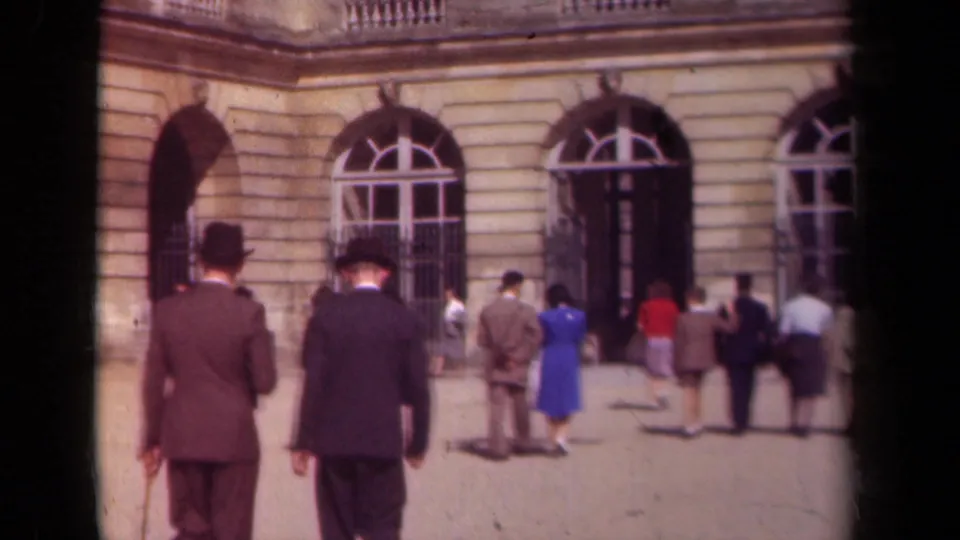 1939 France frame with men in top hats, women in dresses, and other visitors in a sunlit palace-style courtyard.