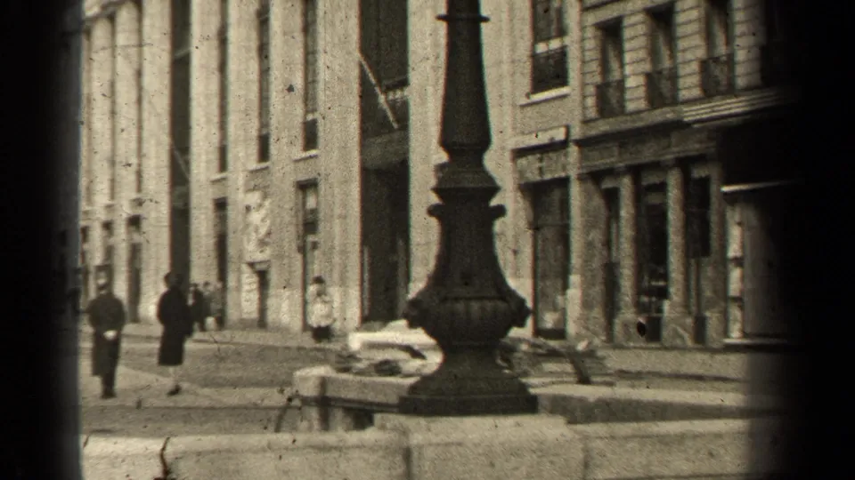 1947 Paris sidewalk scene with a prominent lamppost, pedestrians in dark clothing, stone building fronts, and shop windows.