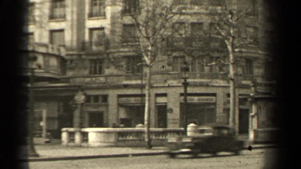 1947 Paris street view with a vintage car passing in front of shopfronts, bare trees, lamppost, and stone railing.