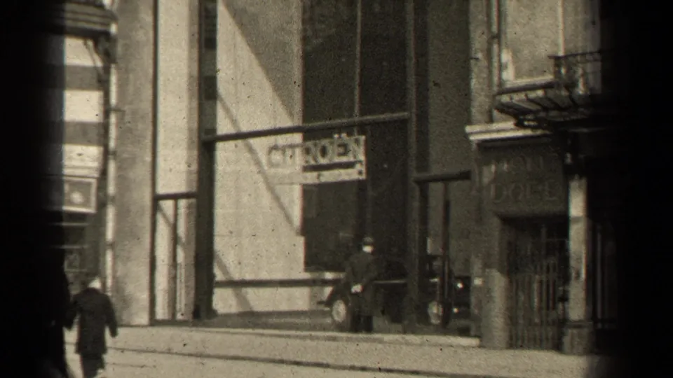 1947 Paris street view with pedestrians in coats moving past storefronts, a Citroen sign, masonry facades, and dark 8mm frame edges.