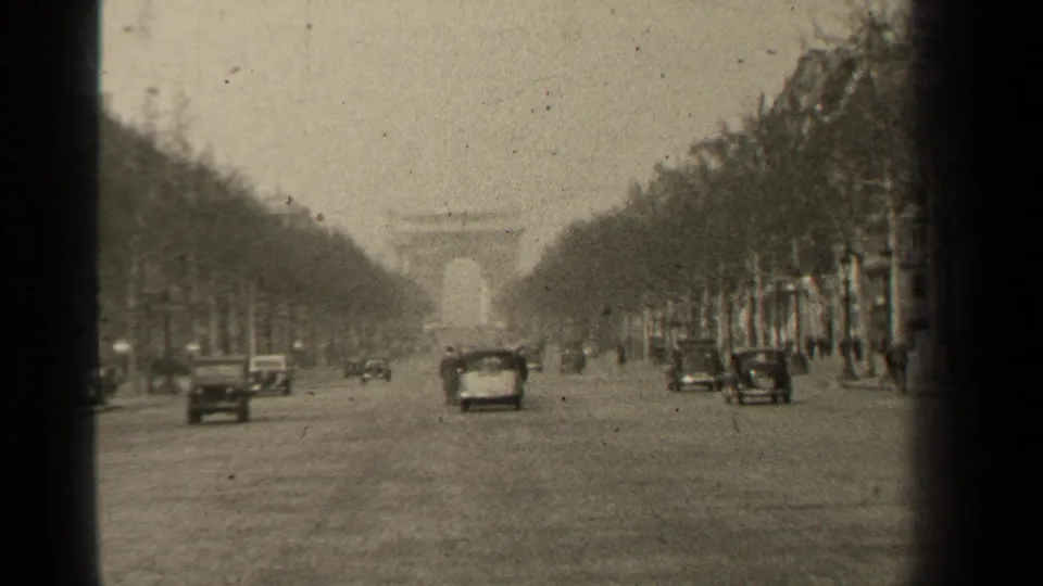 Paris boulevard view with rows of trees, scattered cars, and the Arc de Triomphe visible through haze in the distance.