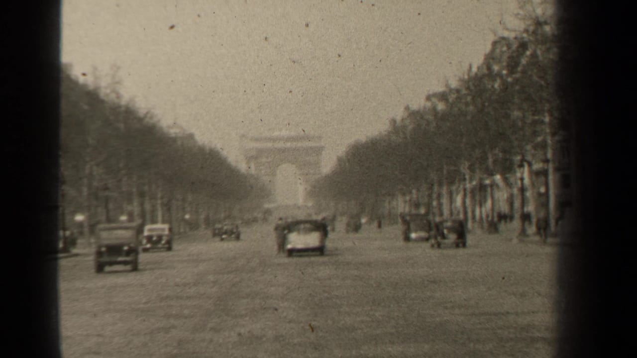 Classic cars travel along a tree-lined Paris boulevard with the Arc de Triomphe in the distance.