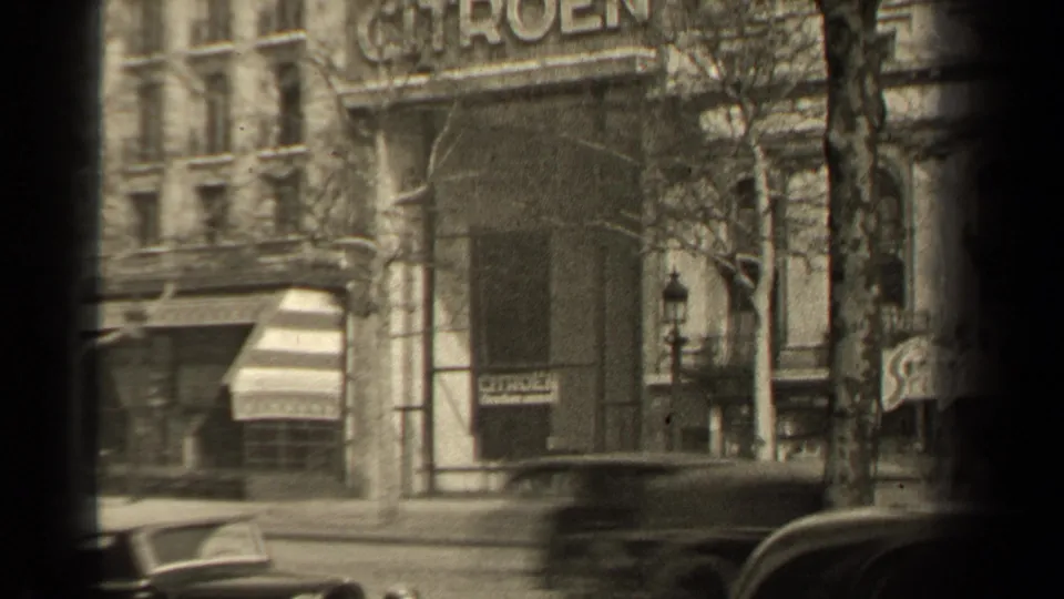 1947 Paris street facade with a large Citroen sign, striped awning, parked cars, bare tree branches, and dark 8mm gate shadow.