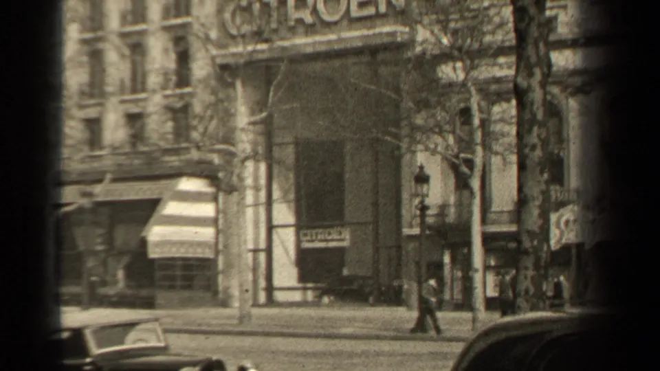 1947 Paris street scene showing a Citroen facade, awning, parked cars, a sidewalk pedestrian, and soft monochrome 8mm texture.