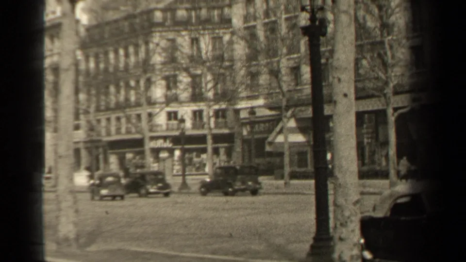 1947 Paris street view showing dark automobiles crossing a broad boulevard beside pedestrians, bare trees, lampposts, and formal building facades.
