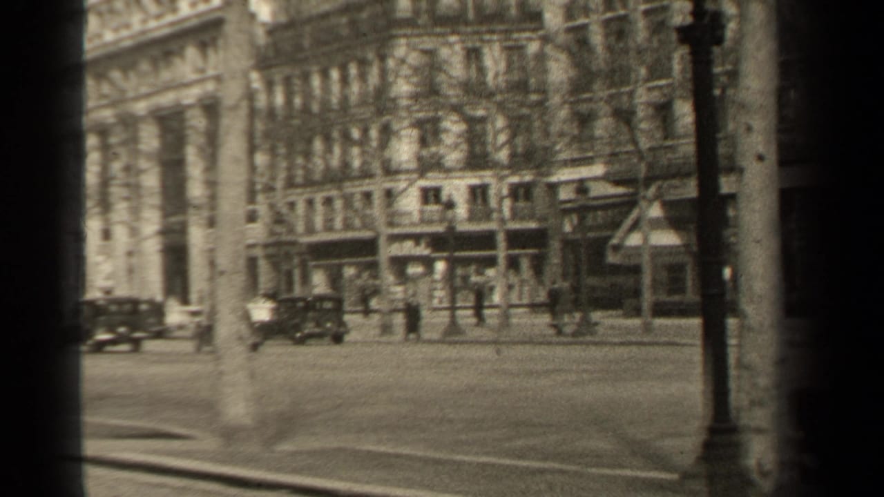 Street corner with parked and moving cars, pedestrians, trees, lampposts, and large buildings.