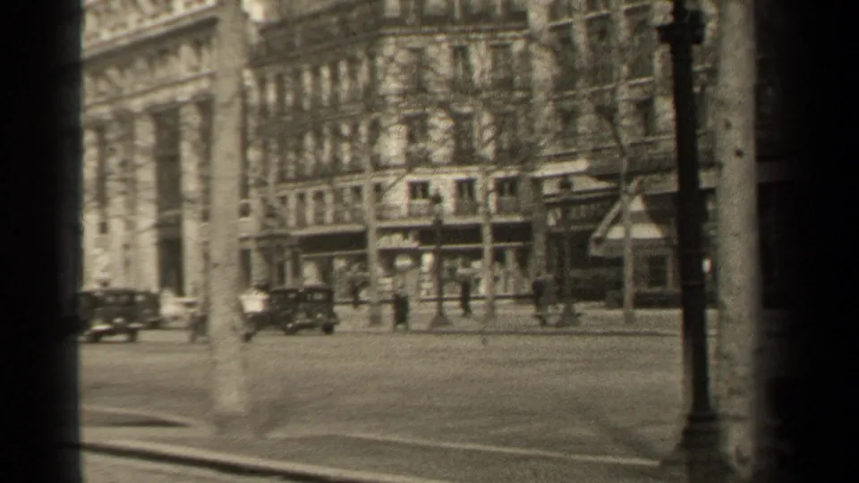 1947 Paris boulevard corner with automobiles near stone-fronted buildings, pedestrians on the far sidewalk, bare trees, and a lamppost in view.
