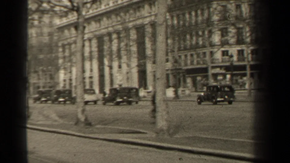 1947 Paris street view with dark cars moving past a curved curb, bare trees, pedestrians, sidewalks, and multi-story stone buildings.