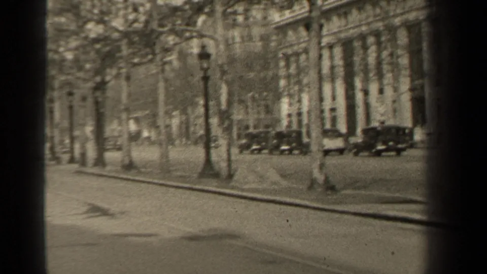 1947 Paris boulevard scene with several automobiles along the street, pedestrians near the far sidewalk, bare trees, lampposts, and stone facades.