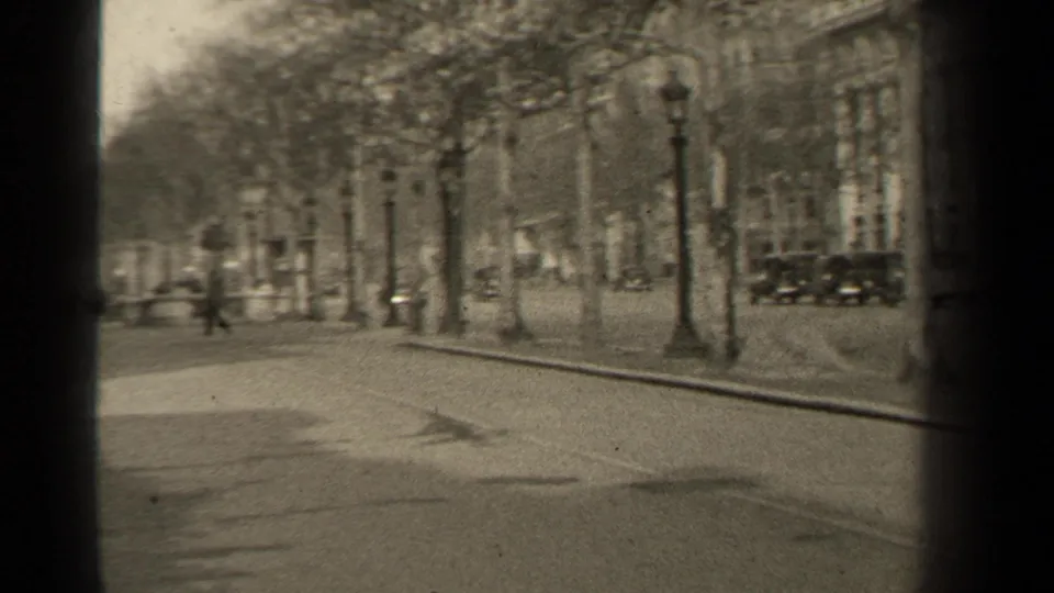 1947 Paris street view along a broad tree-lined boulevard, with pedestrians, vintage cars, lamps, and building facades visible through soft 8mm framing.