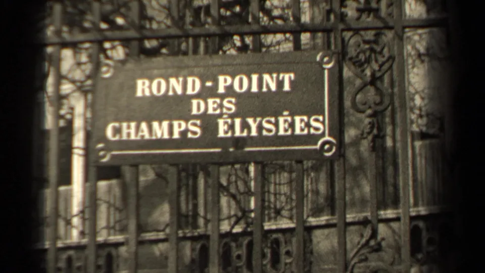 Close view of a Rond-Point des Champs Élysées sign on a metal fence in Paris, with trees and boulevard surroundings behind it.