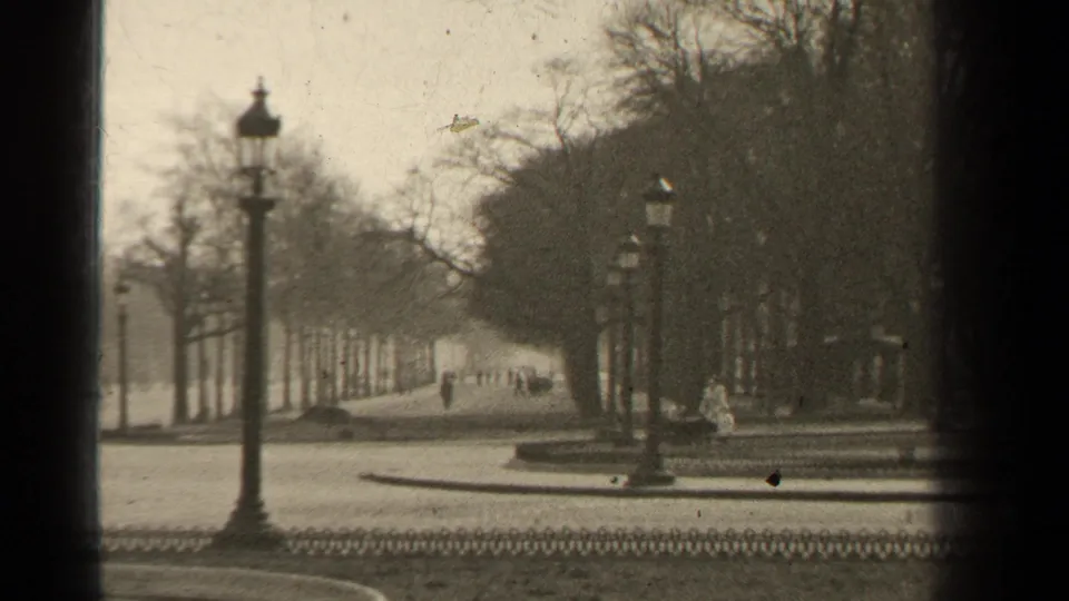 1947 Paris view down a tree-lined street with ornate lampposts, distant pedestrians, low fencing, and soft monochrome 8mm texture.