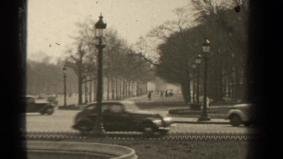 1947 Paris street frame with classic automobiles moving past ornate lampposts, bare trees, low fencing, and distant pedestrians.