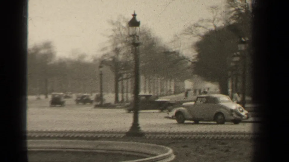 1947 Paris street view with small cars, pedestrians, bare trees, lampposts, and a circular fountain in the foreground.