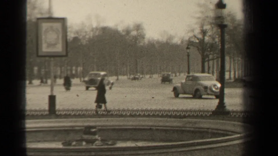 A 1947 Paris boulevard scene shows pedestrians in dark coats, vintage cars, lampposts, and a fountain edge in soft 8mm focus.