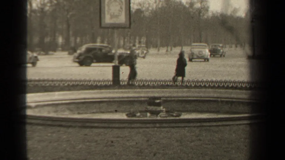 1947 Paris boulevard traffic continues past a circular fountain, with pedestrians in dark clothing and vintage cars under rows of bare trees.