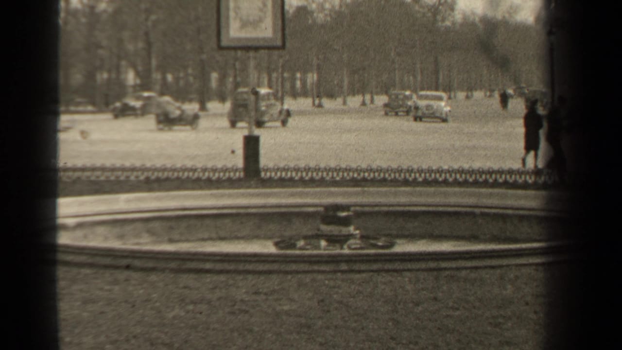 Classic cars pass along a wide boulevard while a pedestrian stands near the right edge.