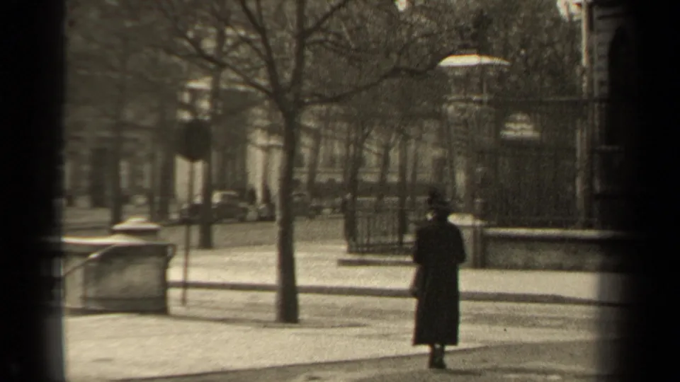 1947 Paris sidewalk view with a woman in a long dark coat near a stone wall, iron gate, bare trees, and street lamps.