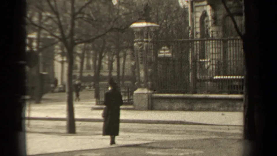 1947 Paris street frame showing a woman in a long coat near iron railings, stone posts, bare trees, and a sidewalk edge.