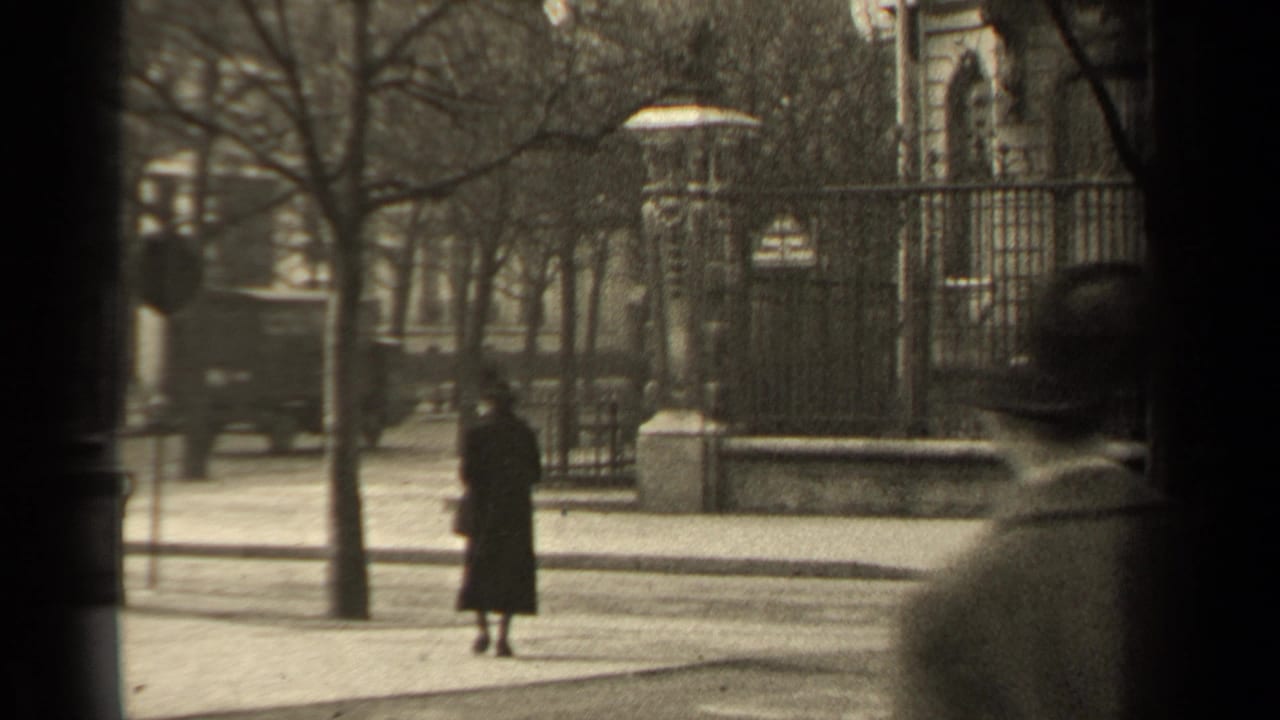 1947 Paris street view with a woman in a long dark coat near wrought-iron fencing, bare trees, pavement, and a car in the background.