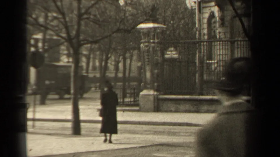 1947 Paris street view with a woman in a long dark coat near wrought-iron fencing, bare trees, pavement, and a car in the background.