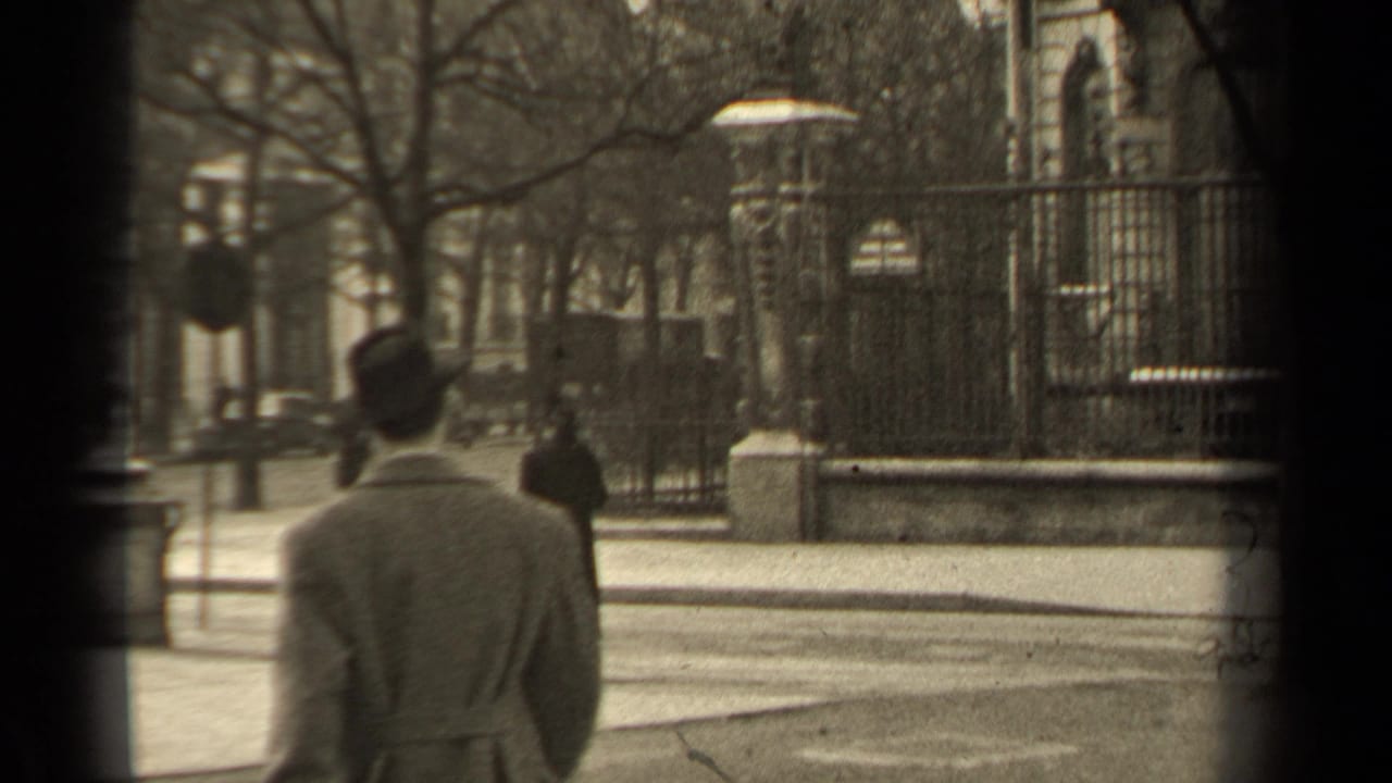Man in a light coat and hat seen from behind near a city street, iron fence, bare trees, and distant pedestrians.