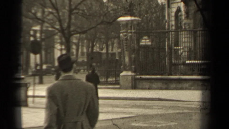 1947 Paris street frame with a man in a light coat seen from behind, a dark-coated pedestrian ahead, iron fencing, and bare trees.