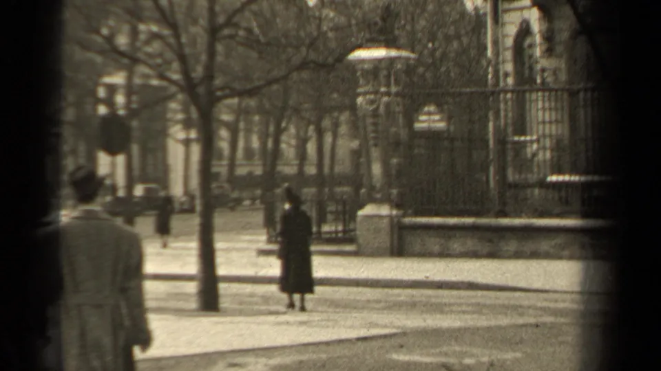 1947 Paris home-movie frame showing a woman in a dark coat near the curb, with a partially seen man, bare trees, and iron fencing.