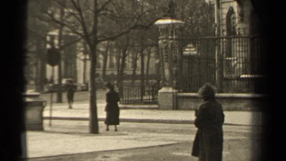 1947 Paris street scene with a woman in a dark coat at right, wrought-iron fencing, bare winter trees, pavement, and distant figures.