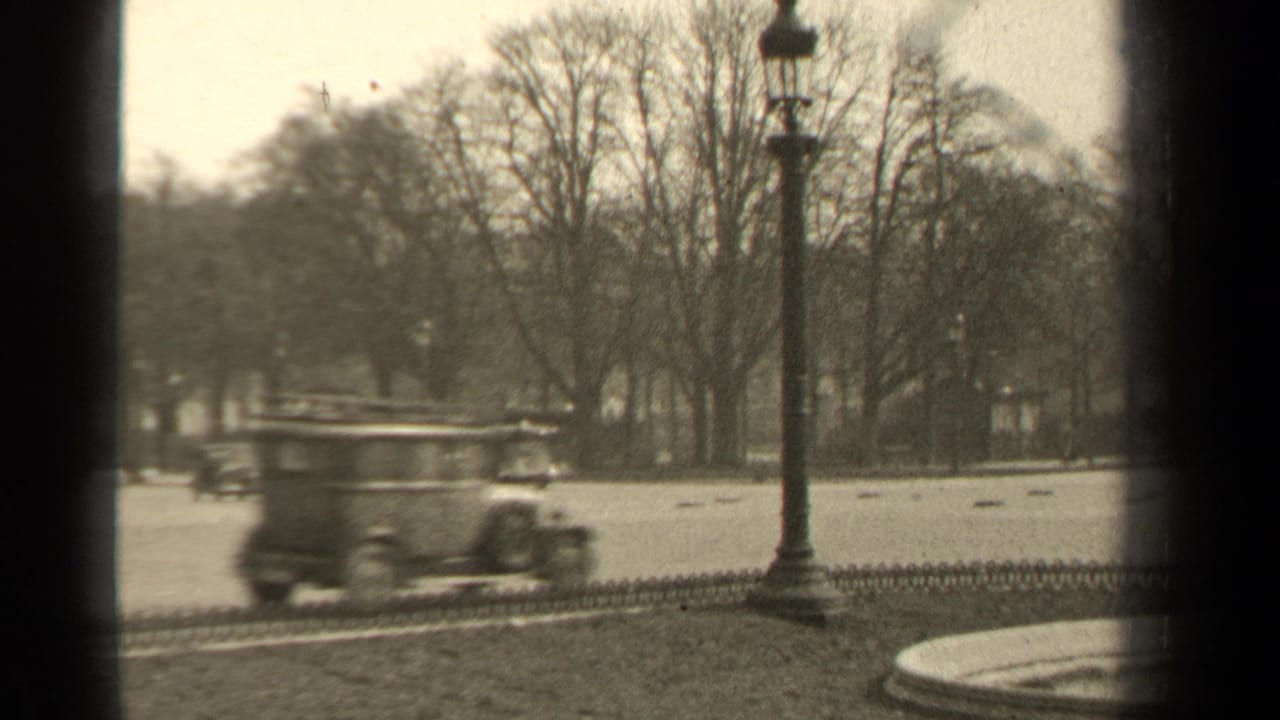 1947 Paris street view with vintage cars moving past bare trees, an iron fence, and a tall streetlamp in soft 8mm footage.