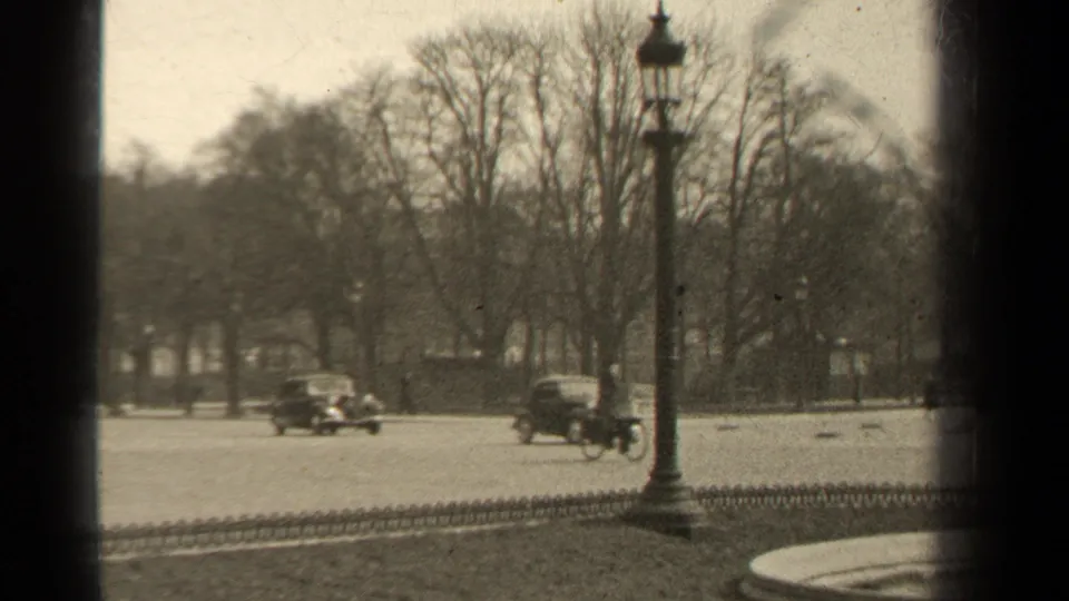 Soft 8mm view of 1947 Paris traffic with two vintage cars, a lamppost, bare trees, and dark edge framing.