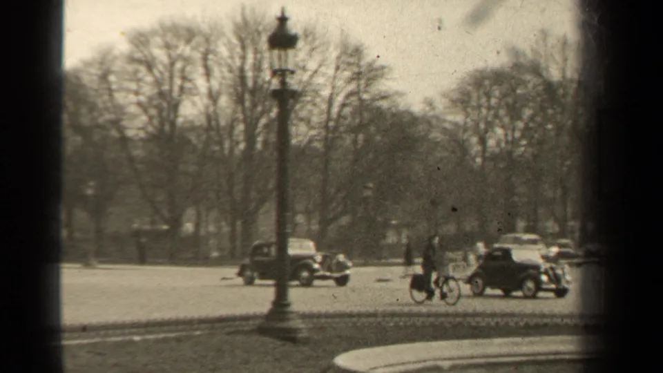 1947 Paris street scene with a cyclist near vintage cars, pedestrians in the distance, bare trees, and an iron fence.