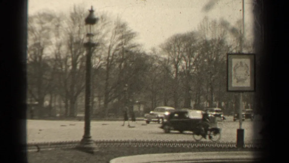 1947 Paris street view with rounded cars, a cyclist, bare trees, iron fencing, and ornate lamp posts in soft 8mm footage.