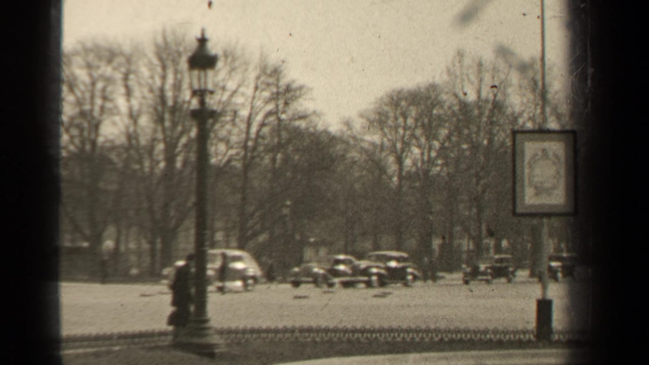 Mid-century cars move along a tree-lined street with a hanging sign and lamp post.