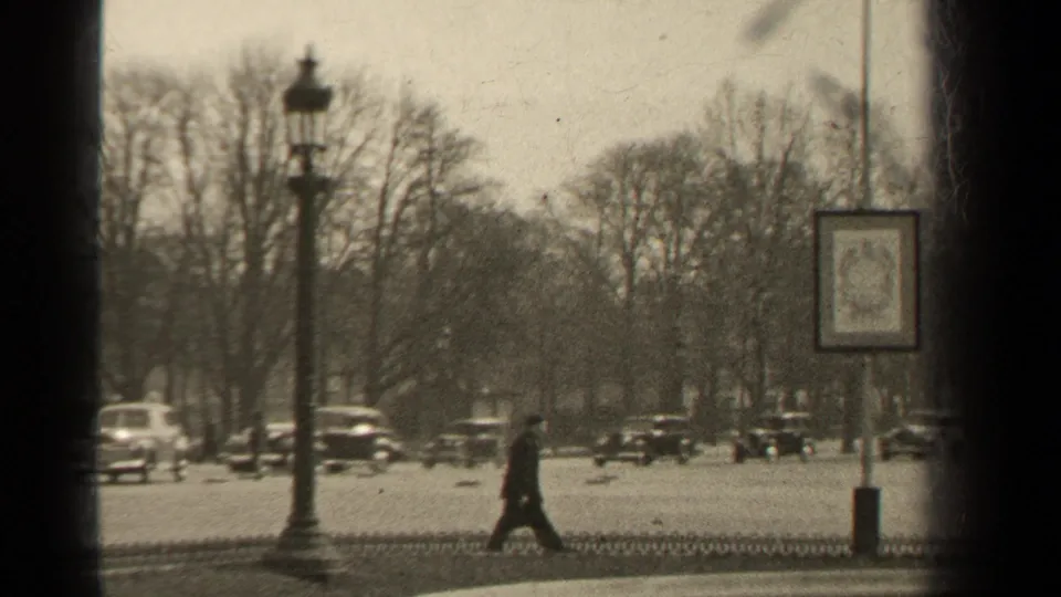 In this 1947 Paris frame, a pedestrian crosses near passing cars, iron fencing, bare trees, a lamp post, and a hanging street sign.