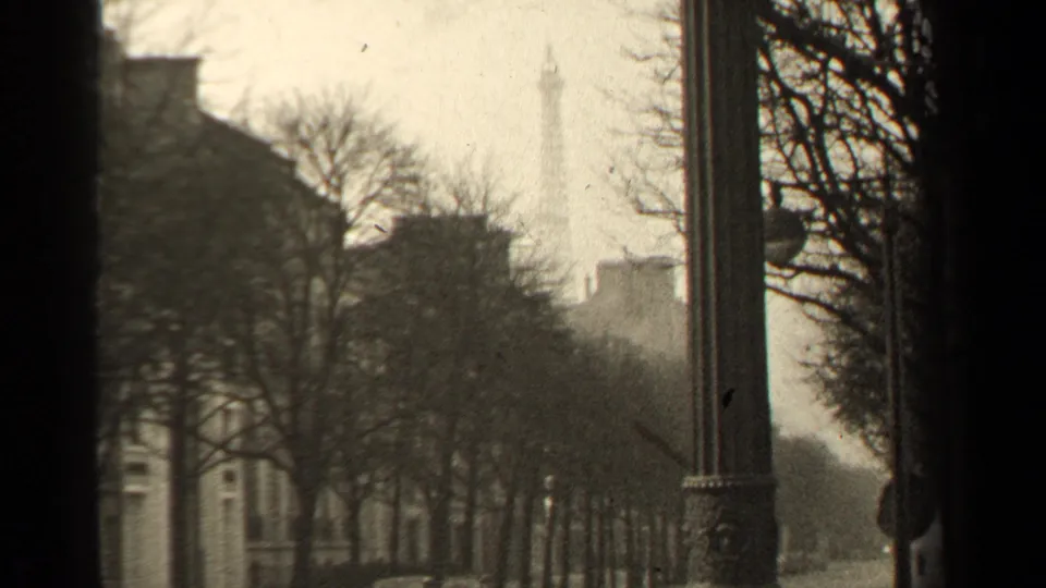 1947 Paris home-movie frame with the Eiffel Tower partly veiled by haze, trees, and foreground street furniture.