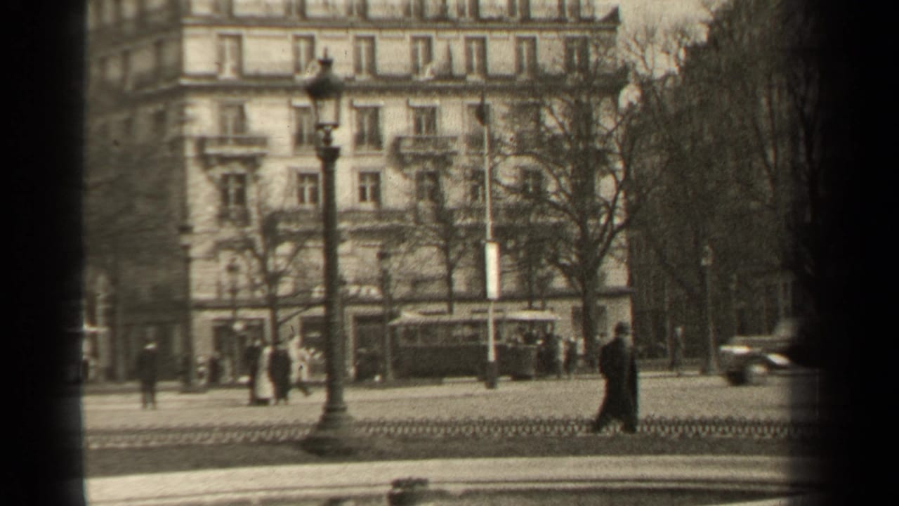 Paris boulevard view showing pedestrians crossing or waiting near lamps, period cars, bare trees, and a stone apartment facade.