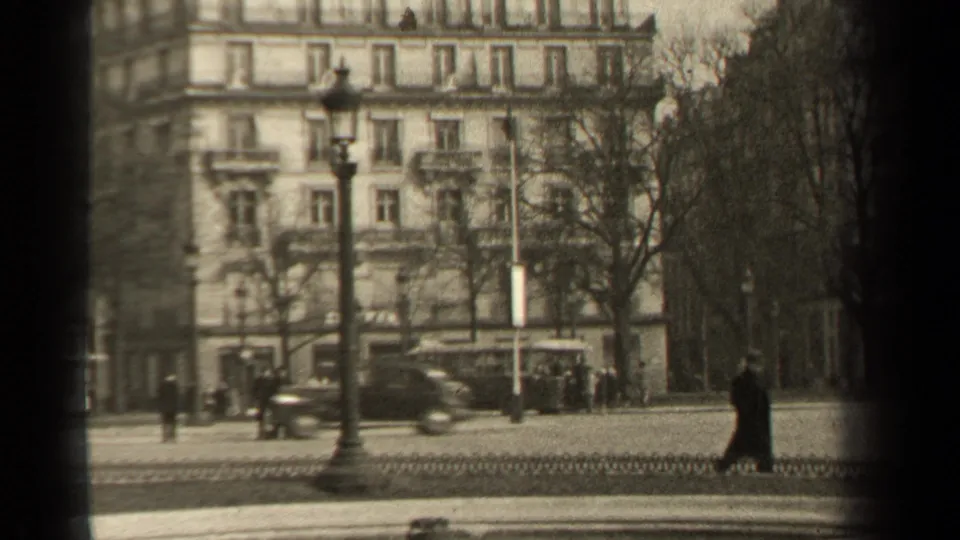 1947 Paris street view with pedestrians in dark coats, a tall lamppost, bare trees, and an ornate apartment facade in soft 8mm focus.