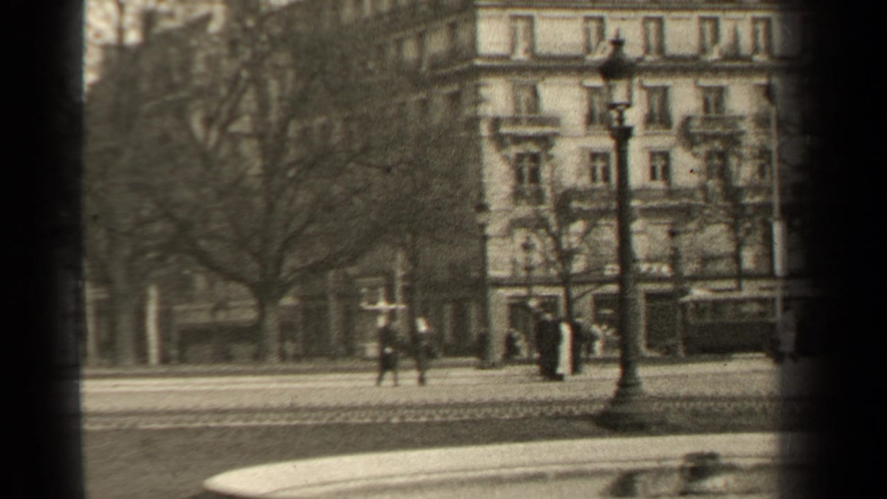 Soft black-and-white 8mm frame of a 1947 Paris street, showing pedestrians, bare trees, a lamppost, and an ornate multi-story facade.
