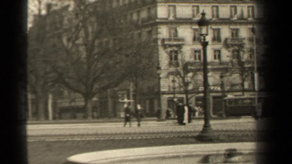 Soft black-and-white 8mm frame of a 1947 Paris street, showing pedestrians, bare trees, a lamppost, and an ornate multi-story facade.