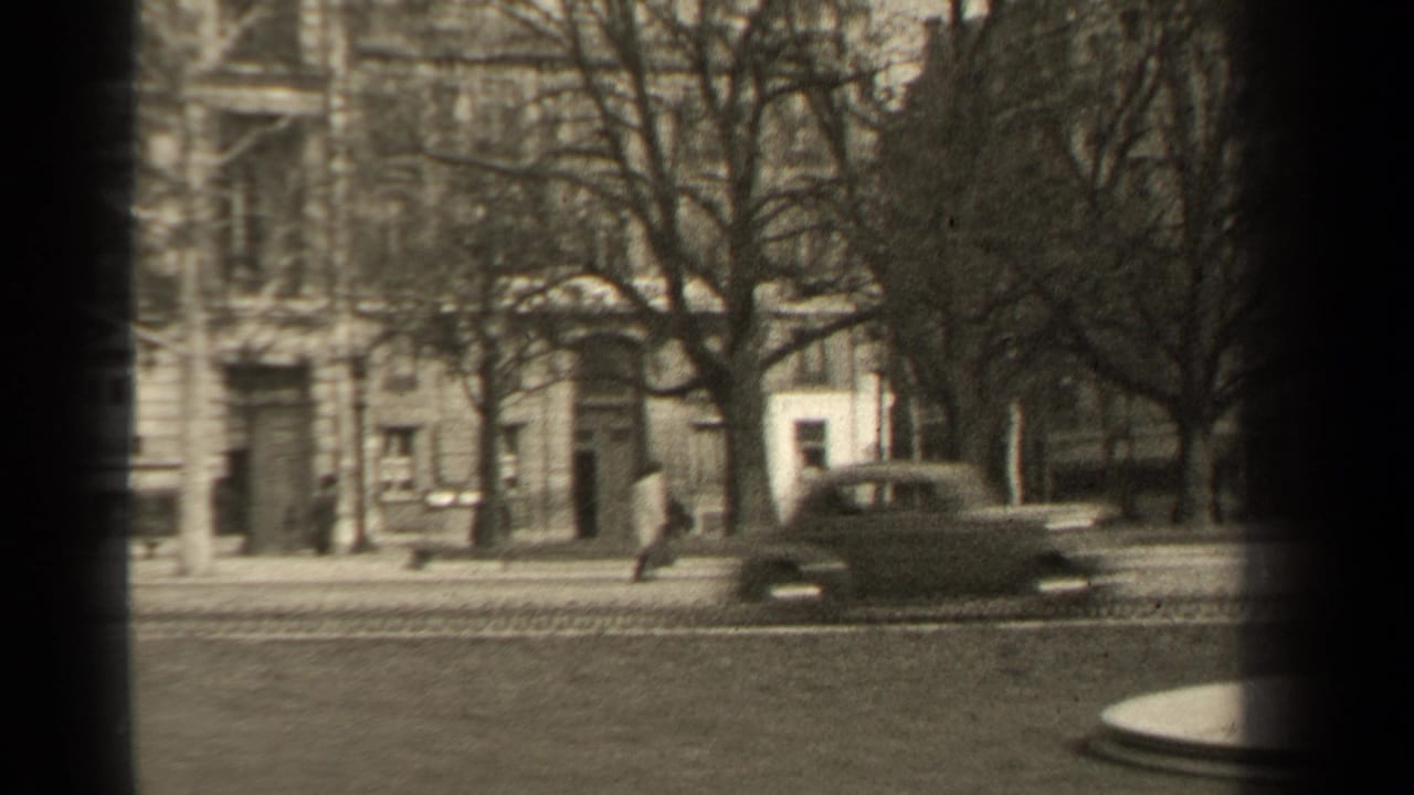 Classic car passes along a tree-lined street in front of masonry buildings and pedestrians.