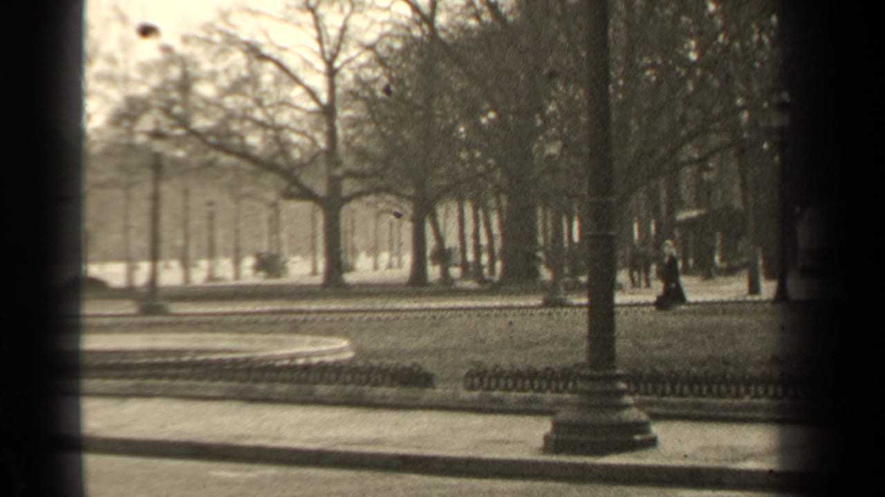 1947 Paris park frame with a woman in dark clothing walking near lampposts, bare trees, railings, and a curving path.