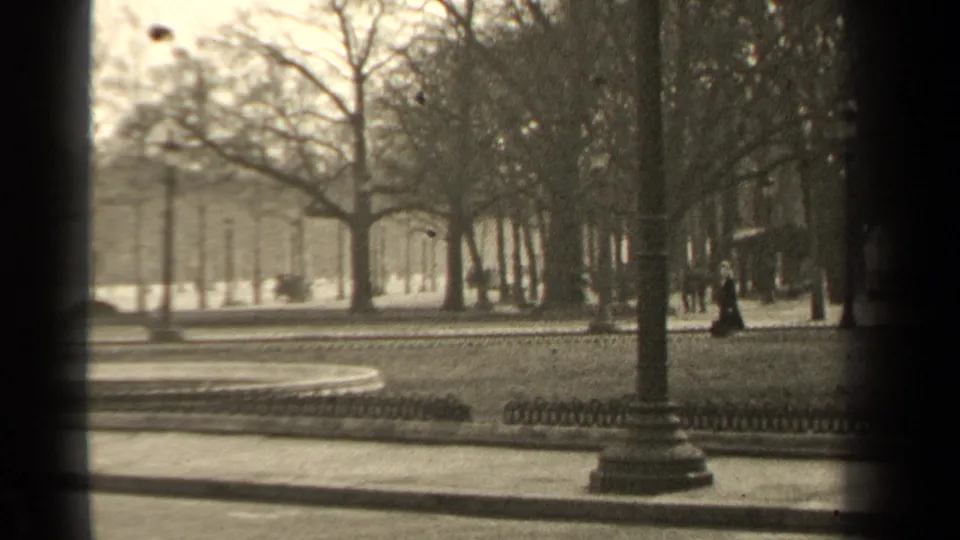 1947 Paris park frame with a woman in dark clothing walking near lampposts, bare trees, railings, and a curving path.