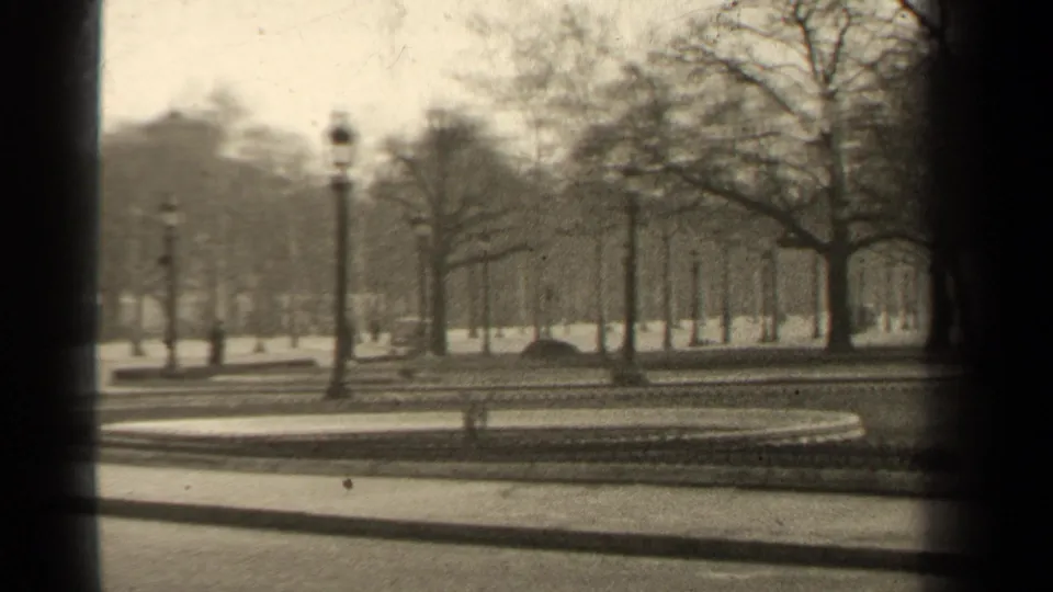 1947 Paris home-movie frame showing a quiet formal park edge with bare trees, ornate lampposts, paths, and a circular bed.