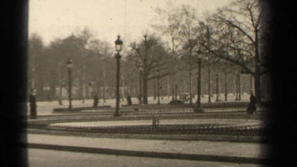 1947 Paris frame with formal park paths, rows of bare trees, ornate lampposts, and a distant pedestrian in a dark coat.